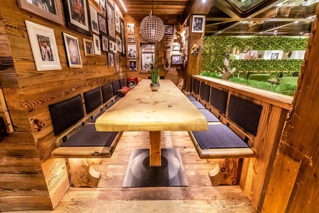 A wooden interior with a long table and benches, framed pictures on the walls, and a hanging light fixture.