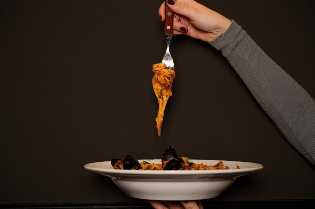 A hand holding a fork with a piece of food above a plate of mussels.