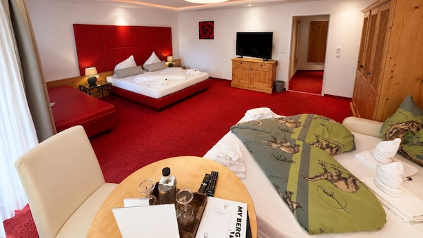 Una habitación de hotel con cama, televisor, armario de madera, sofá rojo, una mesa redonda con bandeja y alfombra roja.