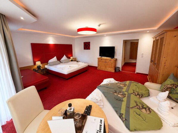 A hotel room with a bed, a TV, a wooden wardrobe, a red sofa, a round table with a tray, and a red carpet.
