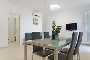 Una sala de estar moderna con una mesa de comedor de vidrio, sillas tapizadas grises, un jarrón azul con una planta, un televisor de pantalla plana sobre un mueble blanco y un cuadro en la pared.