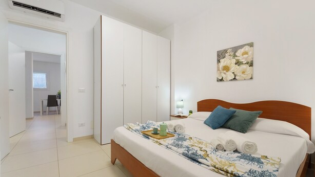 A bedroom with a bed, a bedside table, a nightstand, a painting, and a door leading to another room. - Costa Rei – Delightful Apartment with Garden (Muravera)
