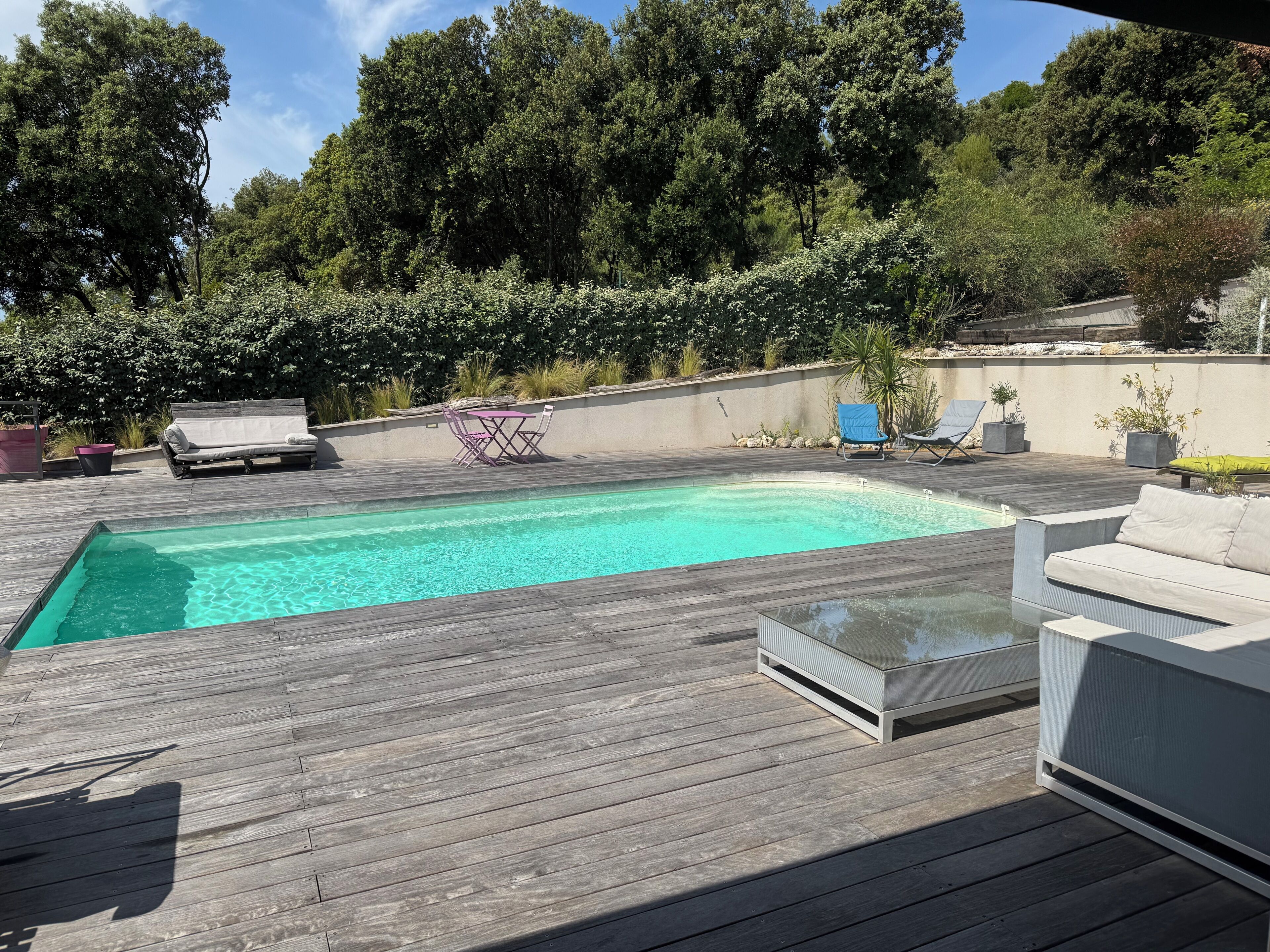 Eine Holzterrasse mit Schwimmbad, Gartenmöbeln und Blick auf Bäume.