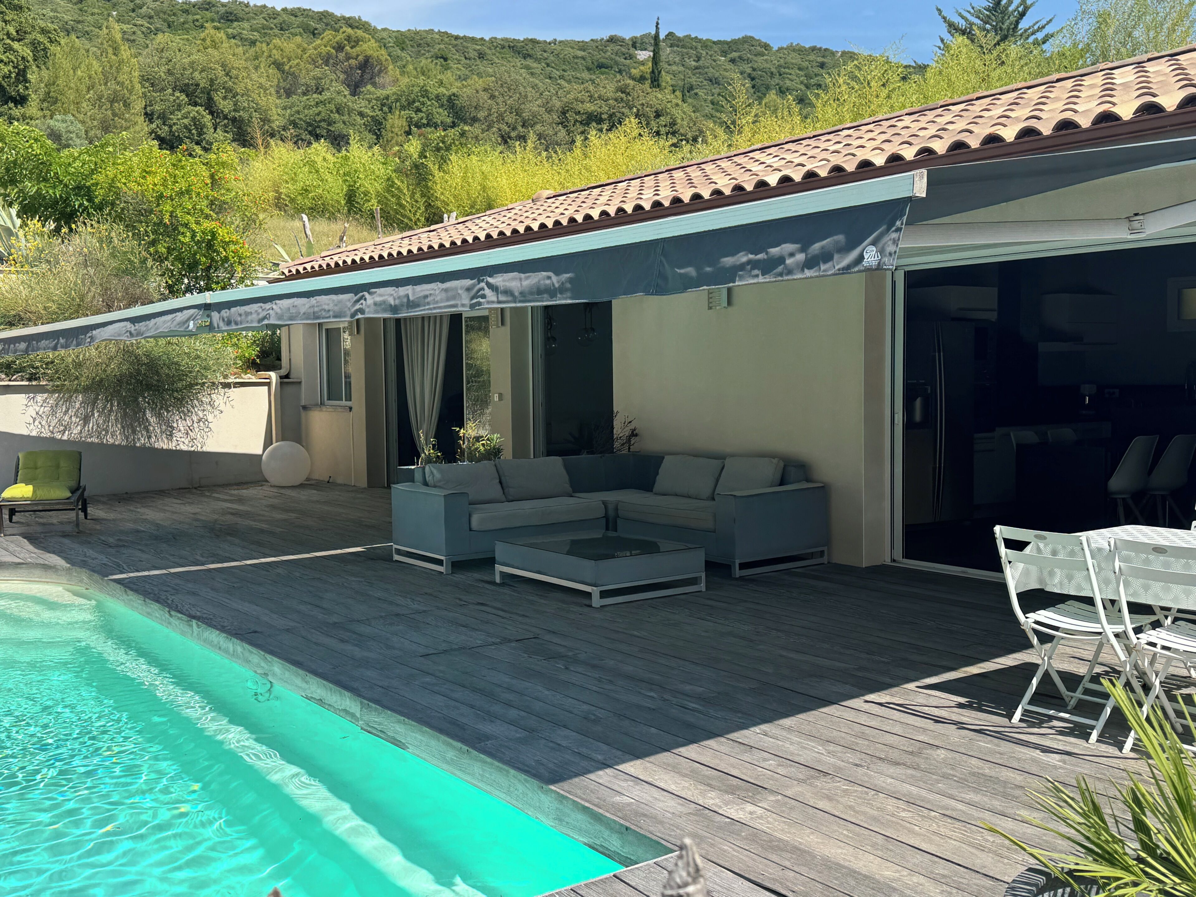 Un espace aménagé au bord de la piscine, comprenant un coin salon couvert, une table à manger avec des chaises et une vue sur la végétation environnante.