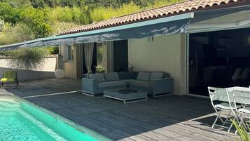 Un espace aménagé au bord de la piscine, comprenant un coin salon couvert, une table à manger avec des chaises et une vue sur la végétation environnante.