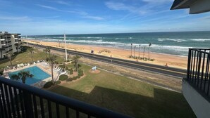 A view from a balcony overlooking a beach with a swimming pool, a road, and a building in the background. - 2-Bedroom 2-Bathroom 5th "TOP" Floor Condo in Stunning Ormond Beach with AC (Ormond Beach)