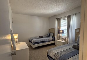 A bedroom with two beds, a bedside table with a lamp, a nightstand, and a window with curtains. - Gorgeous Ocean & Pool View 5th "TOP" Floor 2/2 Condo in Beautiful Ormond Beach (Ormond Beach)