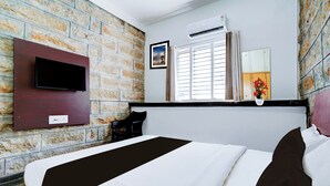 A hotel room with a brick wall, a mounted TV, a window with blinds, a bedside table with a vase, and a bed with white and black bedding. - Super Collection O Rajkumar Road (Mysore)