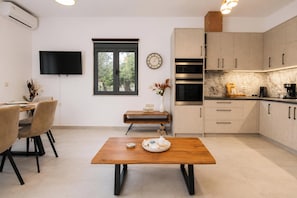 A modern kitchen with a built-in oven, a dining area with a wooden table, and a TV mounted on the wall. - Villa Mark (Glossa)