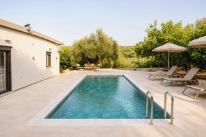 A rectangular swimming pool with a clear blue water surface, surrounded by a tiled deck. There are lounge chairs and a table with a canopy, and a stone wall in the background. - Villa Mark (Glossa)