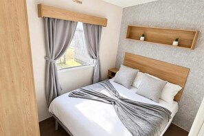 A bedroom with a wooden headboard, a bed with white bedding, grey curtains, and a small shelf above the bed. - Coastal Caravan at Lyons Robin Hood, Rhyl (Rhyl)