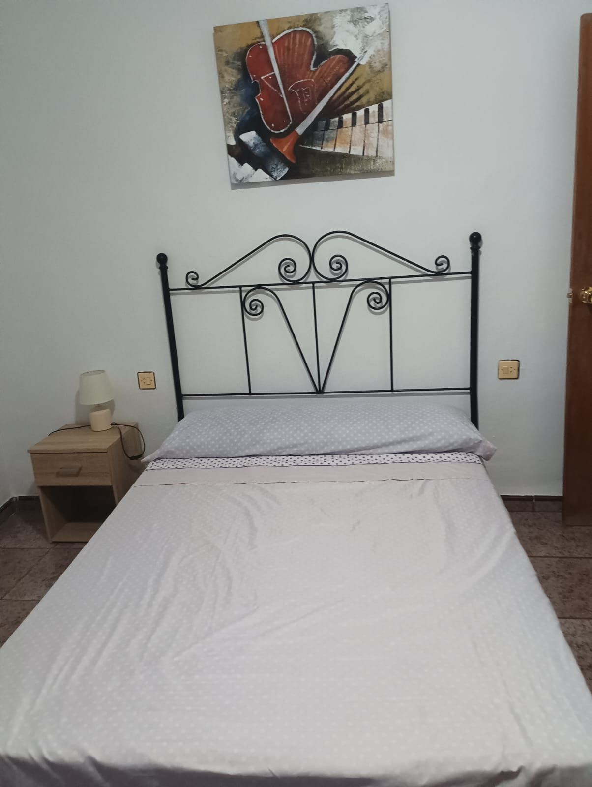Een eenpersoonsbed met een metalen hoofdbord, een nachtkastje met een lamp en een aan de muur hangend schilderij.