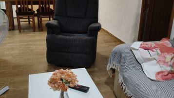 A living room with a sofa, a coffee table with a vase of flowers, a remote control, and a bed with a blanket.