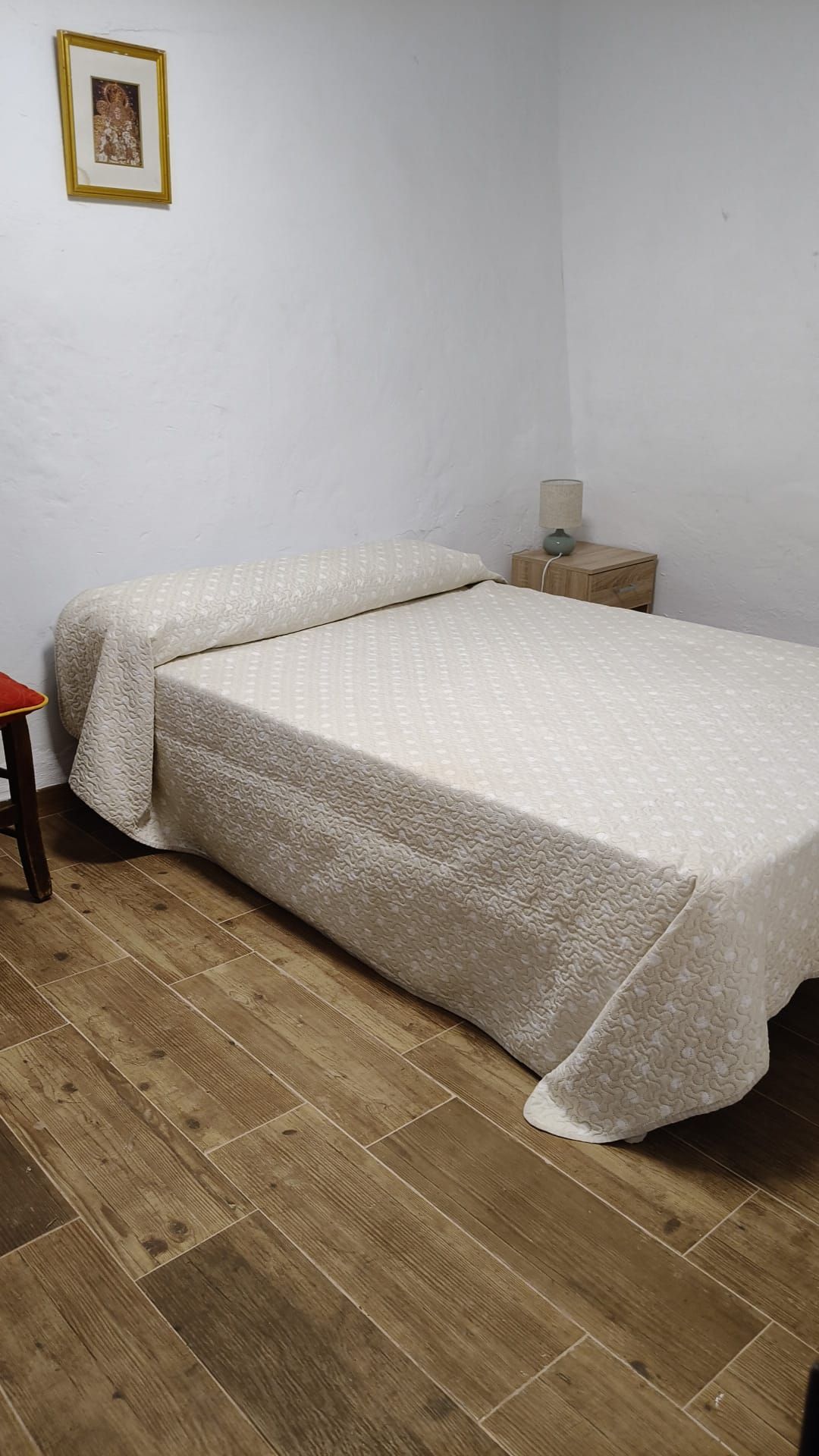 A small, single-bed room with a wooden floor, a bedside table, and a lamp.