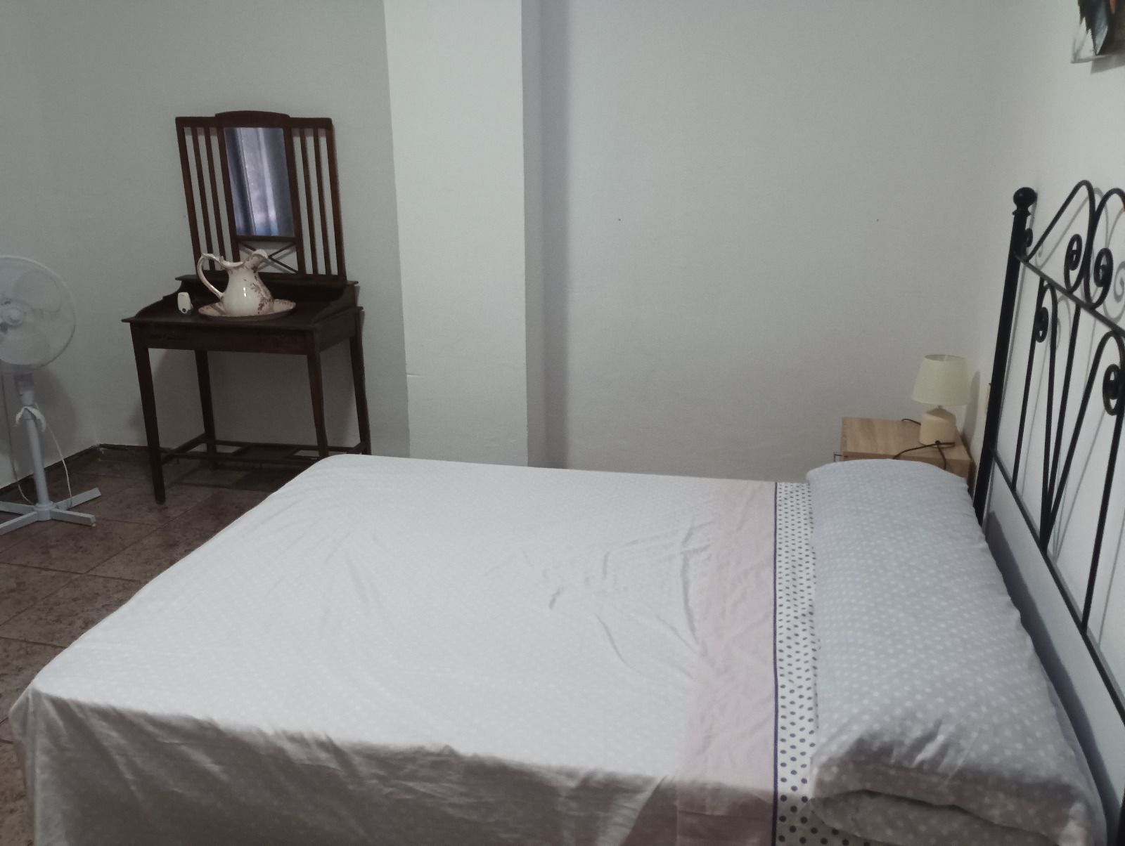 Quarto com cama, mesa de cabeceira, cadeira, espelho e ventilador.