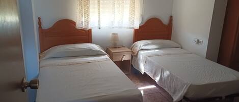 A room with two single beds, a nightstand with a lamp, a window with lace curtains, and a door with a brass handle.