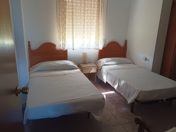 Una habitación con dos camas individuales, una mesita de noche con lámpara, una ventana con cortinas de encaje y una puerta con picaporte de latón.