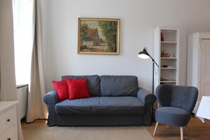 A living room with a grey sofa, a red pillow, a blue armchair, a painting on the wall, and a white cabinet.