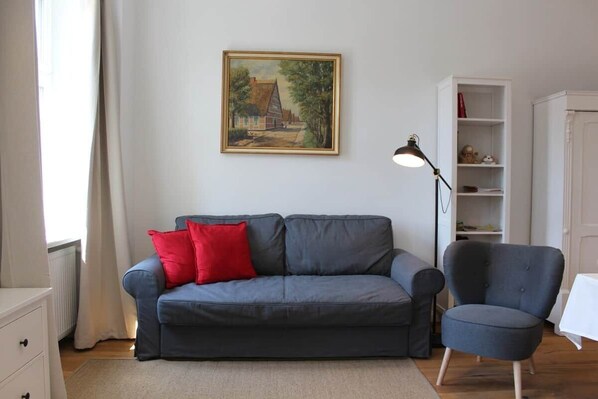 A living room with a grey sofa, a red pillow, a blue armchair, a painting on the wall, and a white cabinet.