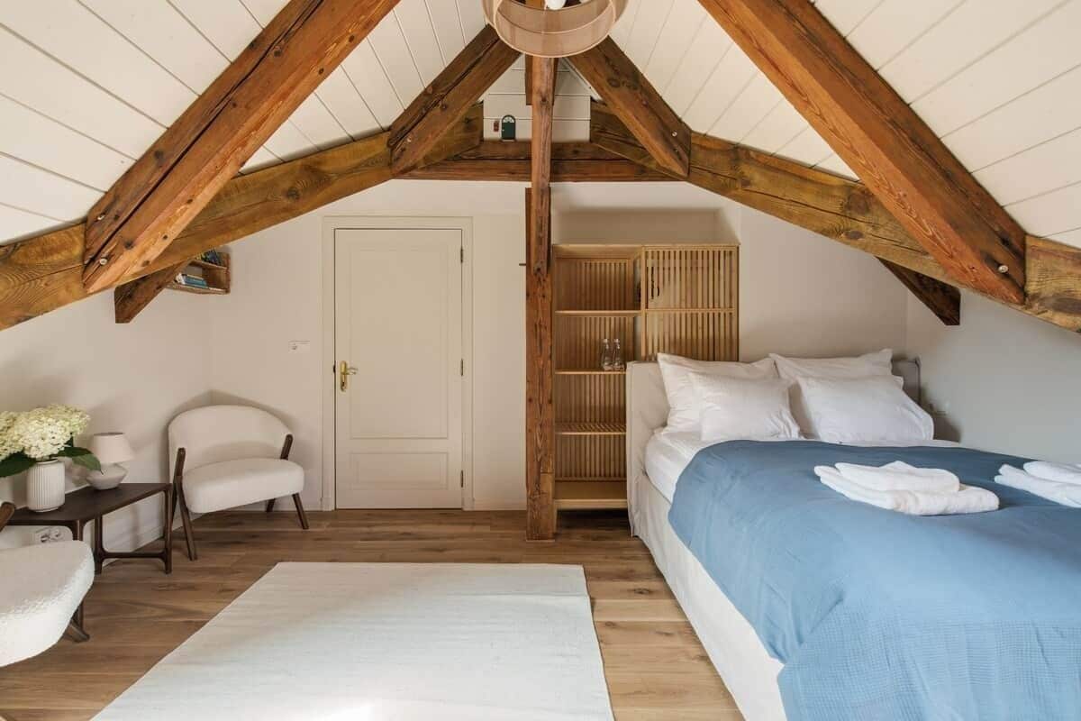 Een slaapkamer met een schuine kap, een bed met een blauwe spreideken, een witte stoel, een nachtkastje met een lamp, en een houten garderobe.