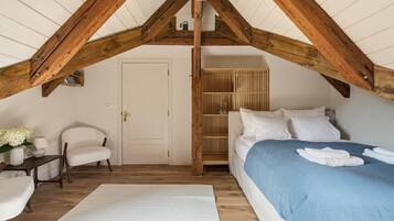 Een slaapkamer met een schuine kap, een bed met een blauwe spreideken, een witte stoel, een nachtkastje met een lamp, en een houten garderobe.