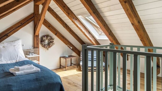 A room with a sloped ceiling, wooden beams, a bed with a blue cover, a small table with a lamp, and a crib.