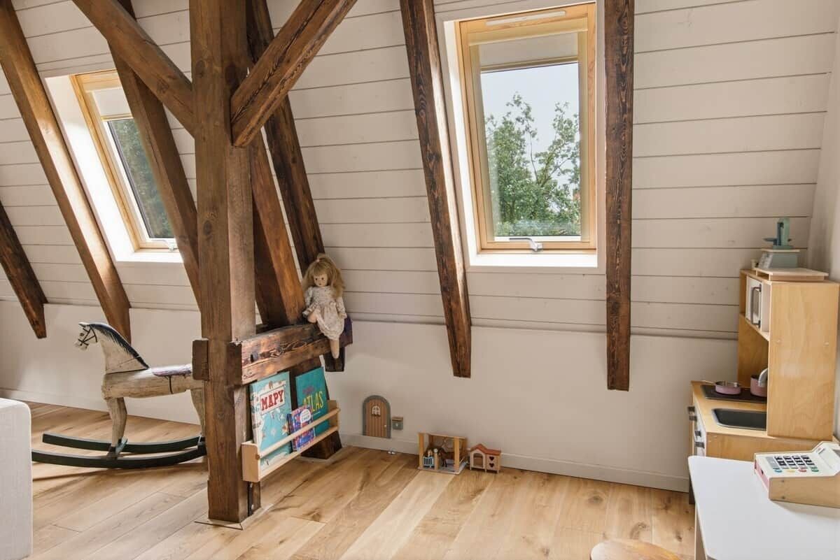 A room with wooden beams, a rocking horse, a small shelf with toys, and a window with a view of trees.