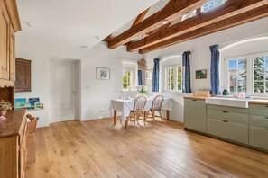 Eine geräumige Küche mit Holzbalken, ein Essbereich mit Tisch und Stühlen sowie ein Blick ins Freie durch große Fenster.