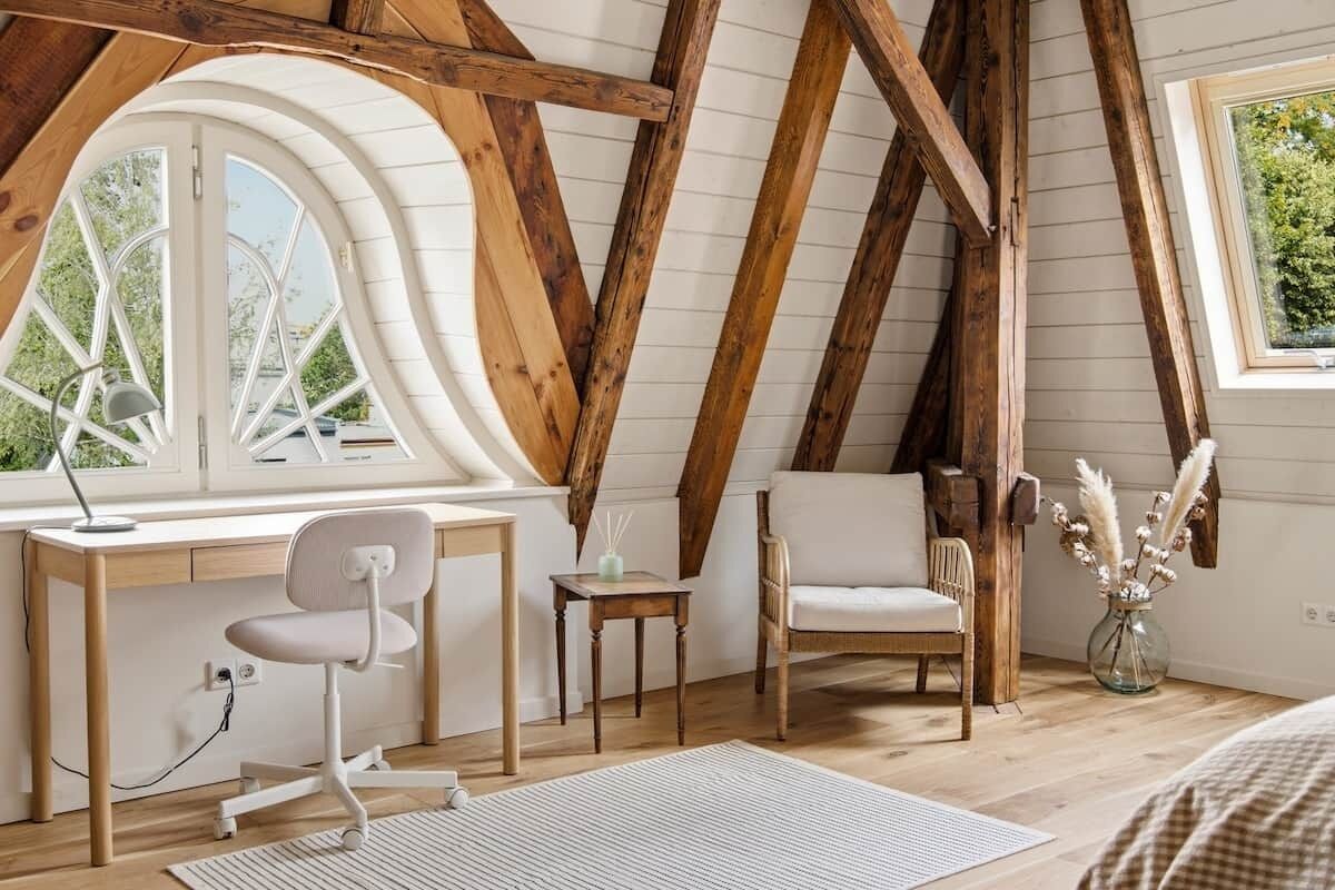 Ein Zimmer mit schrägem Dach, Holzbalken, einem Schreibtisch mit Stuhl, einem Fenster und einem kleinen Tisch mit einer Vase getrockneter Pflanzen.