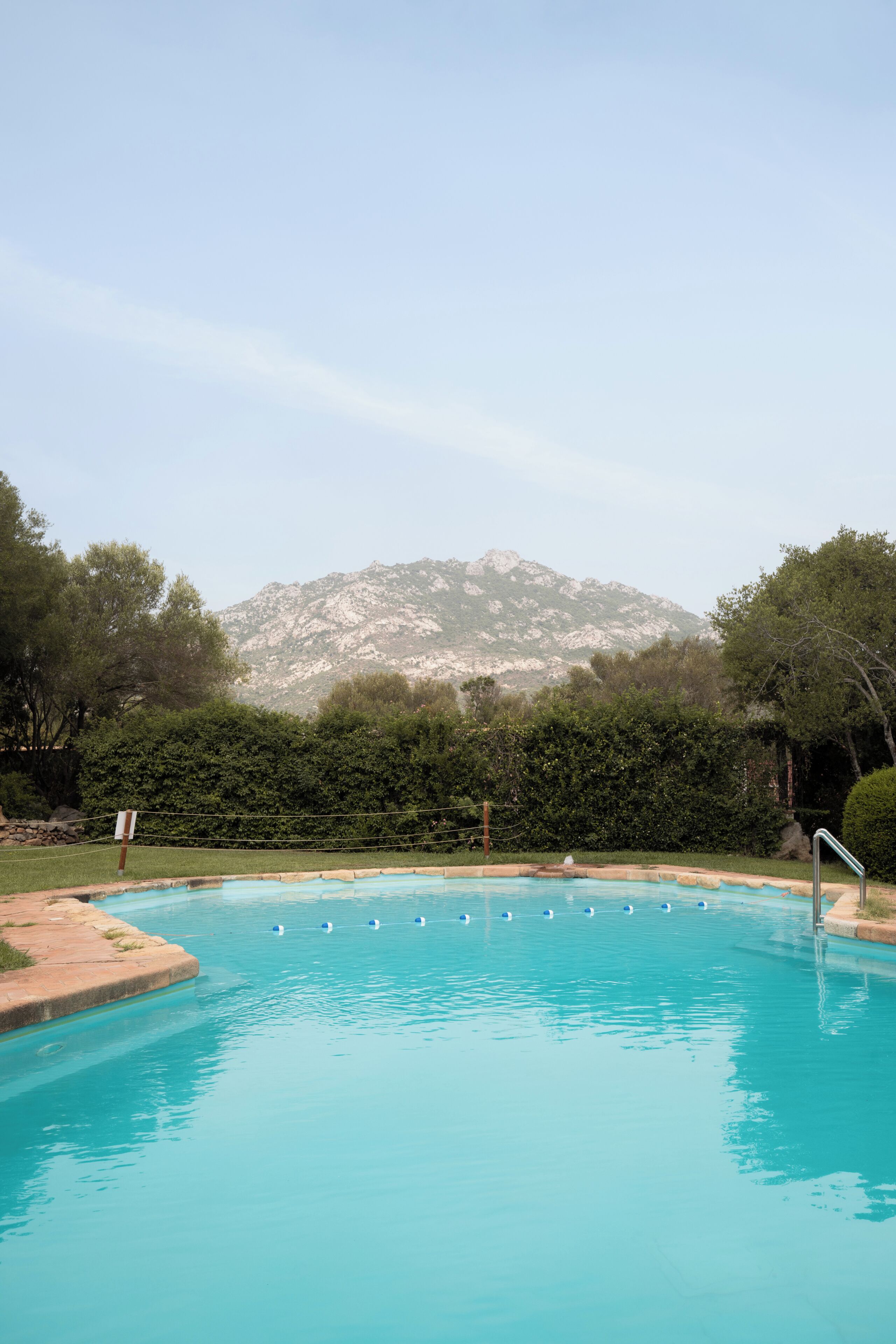 A swimming pool with clear blue water, surrounded by greenery and a mountain in the background.