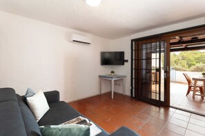 Sala de estar com sofá, uma mesinha de centro com uma planta, televisão de tela plana e uma porta de vidro deslizante que dá para uma área externa com mesa e cadeiras.