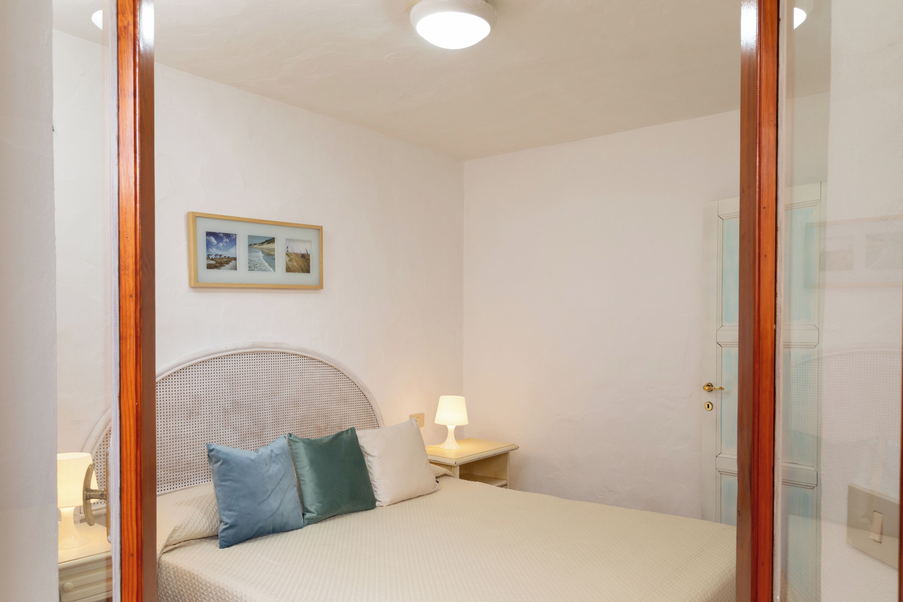 A bedroom with a bed, a bedside table with a lamp, a wall-mounted picture, and a door.