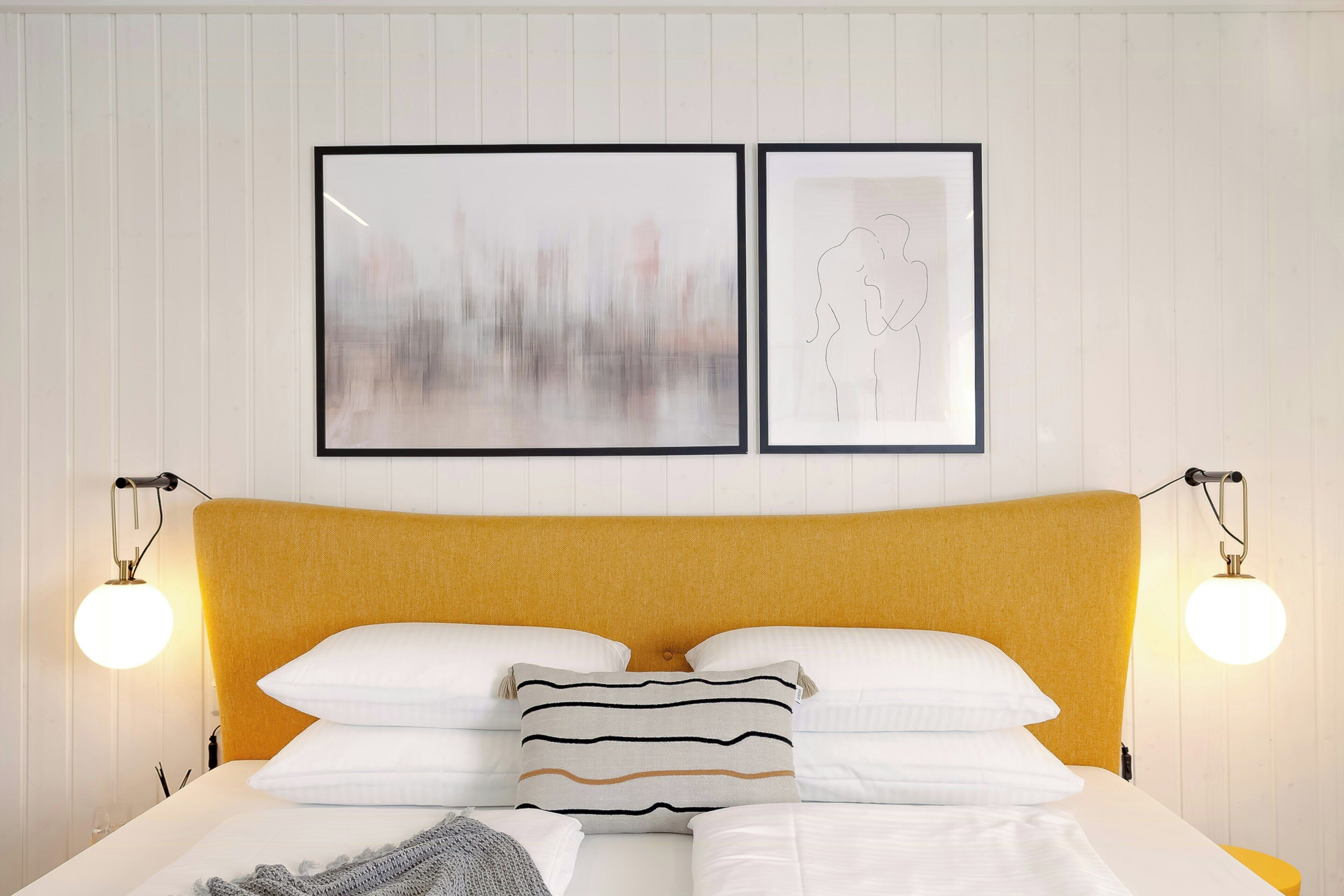Una cama con cabecera amarilla, ropa de cama blanca y dos lámparas fijadas a la pared.
