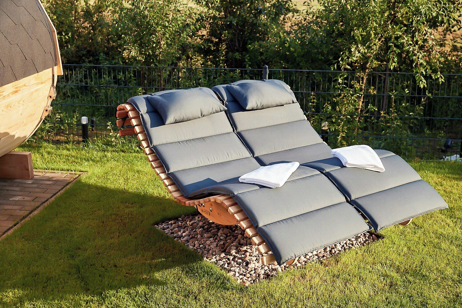 A lounge chair with a cushion and a towel placed on it, set on a grassy area with a brick structure in the background.