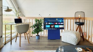 A modern living room with a flat-screen TV, a laptop, a potted plant, a wooden chair, a white pouf, and a glass table with a remote control. - Design retreat with sauna near Berlin (Wandlitz)