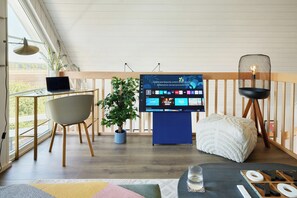 Ein modernes Wohnzimmer mit Flachbildfernseher, Laptop, Topfpflanze, Holzstuhl, weißem Pouf und einem Glastisch mit Fernbedienung.