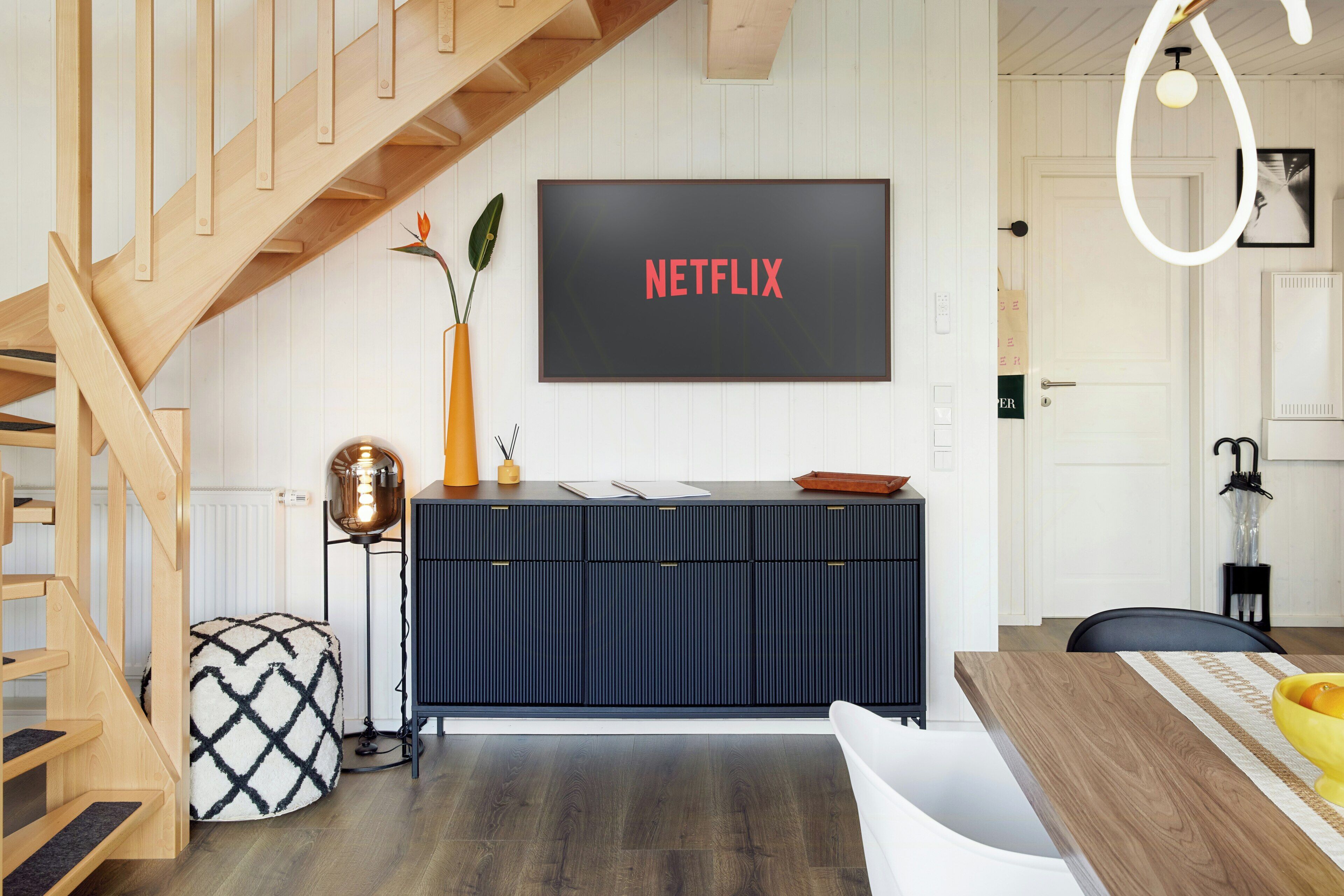 Ein modernes Interieur mit einer Holztreppe, einem schwarzen Rahmen um den Fernseher, auf dem 'NETFLIX' angezeigt wird, und einem Essbereich mit einem Holztisch und Stühlen.