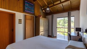 A bedroom with a large bed, wooden wardrobe, a desk with a chair, and a view through a glass door.