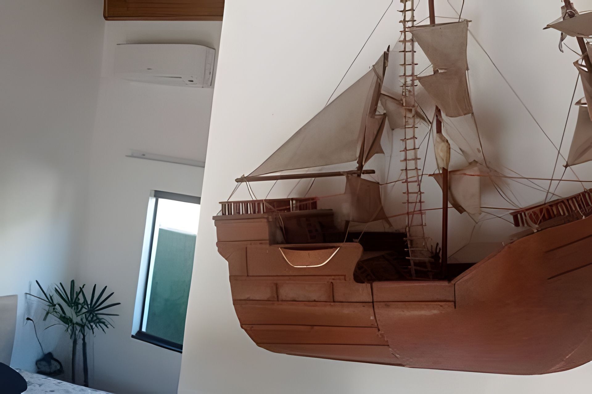 A model ship suspended from the ceiling in a room with a window and a plant.