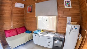 A compact room with a bed, a small kitchenette, a window with a blind, and a wooden ceiling.