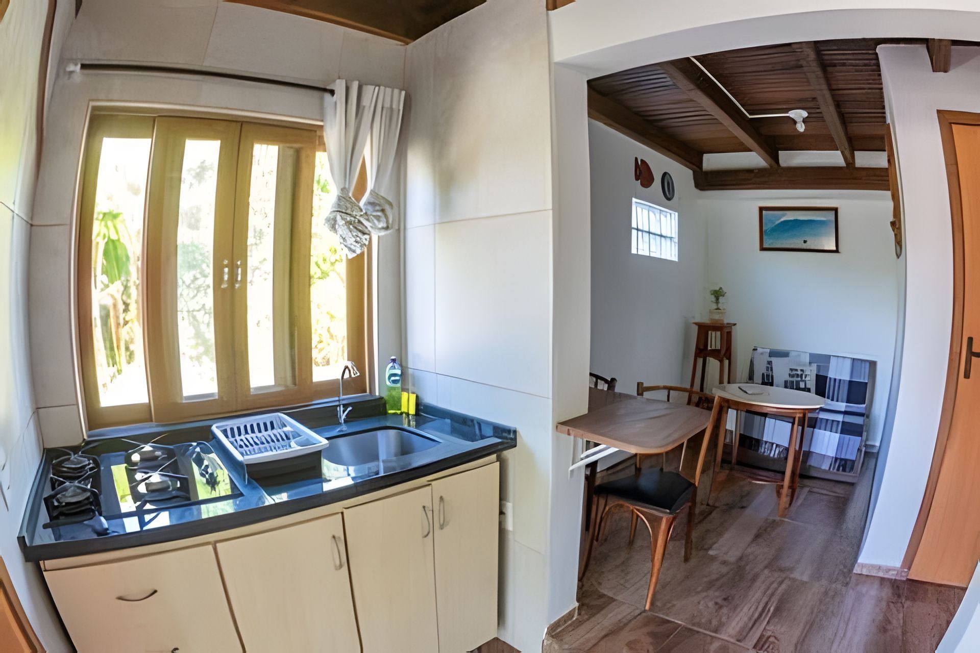 Cozinha com pia, armários e janela com cortinas. Há uma área de refeições com mesa e cadeiras, e vista para o exterior.