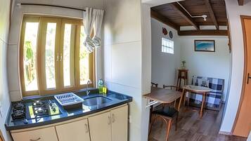 Cozinha com pia, armários e janela com cortinas. Há uma área de refeições com mesa e cadeiras, e vista para o exterior.