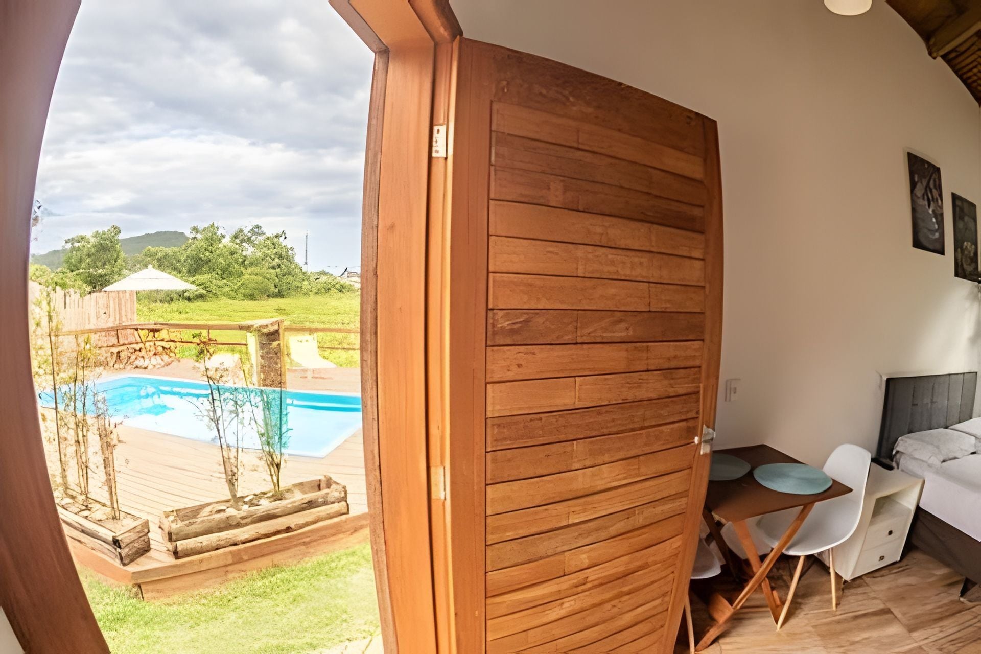 Uma porta de madeira dá acesso a uma área de piscina com mesa e cadeiras, e um pequeno sofá branco dentro de um quarto.