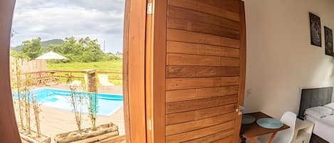 Uma porta de madeira dá acesso a uma área de piscina com mesa e cadeiras, e um pequeno sofá branco dentro de um quarto.