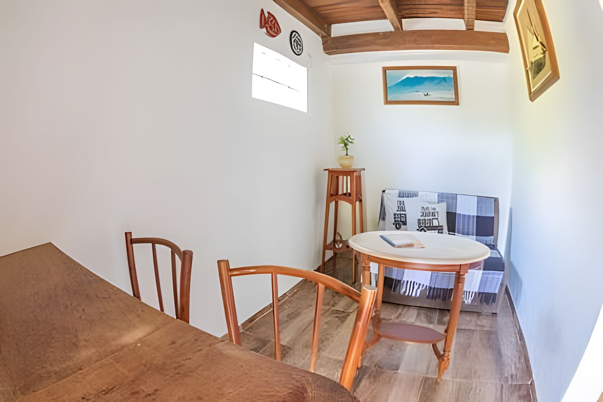Un comedor con una mesa y sillas de madera, una pequeña mesa redonda con una sola silla, una planta en maceta y un cuadro en la pared.