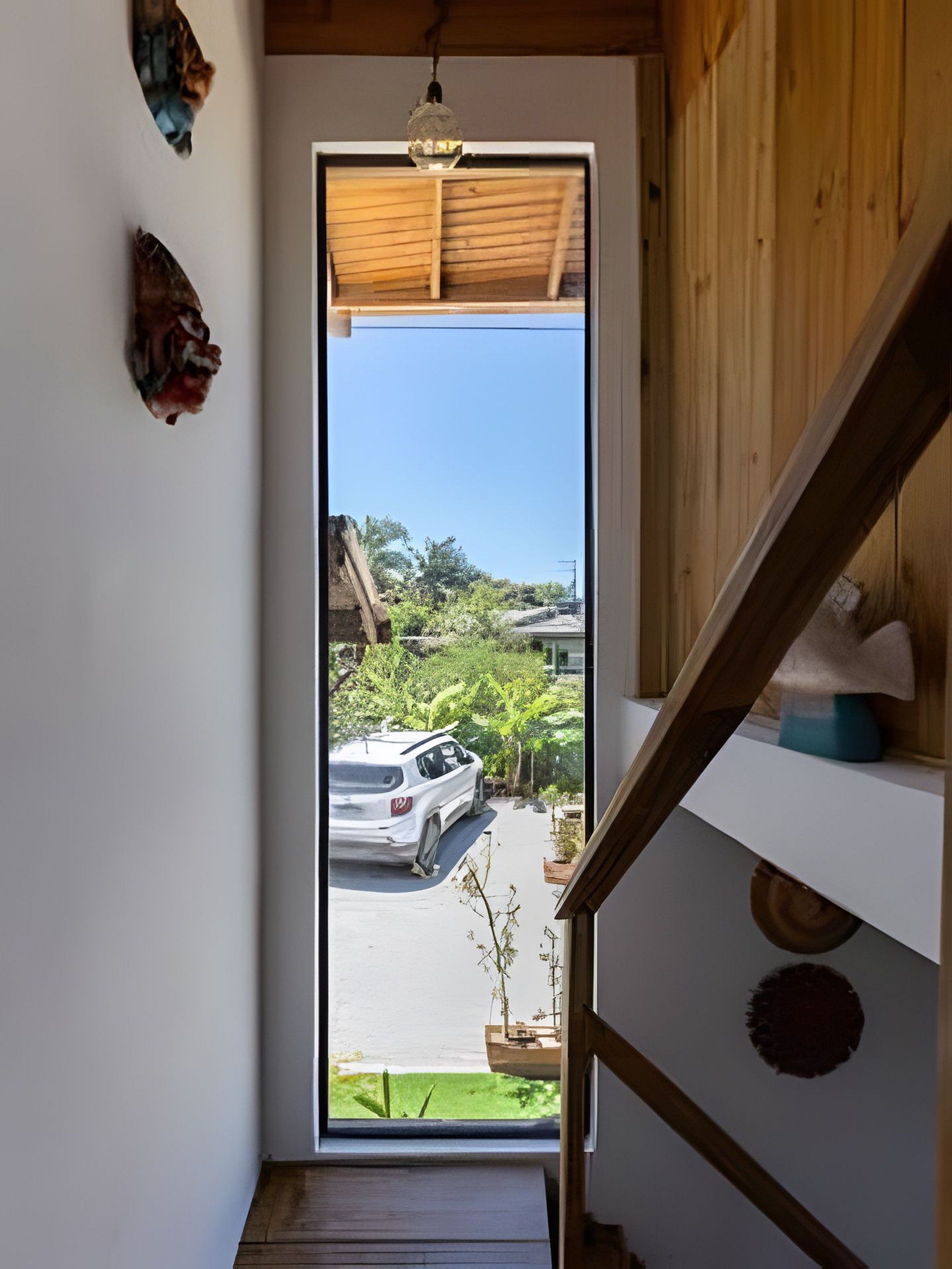 A view through a door to a street with a parked car, a wooden staircase, and a hanging light fixture.