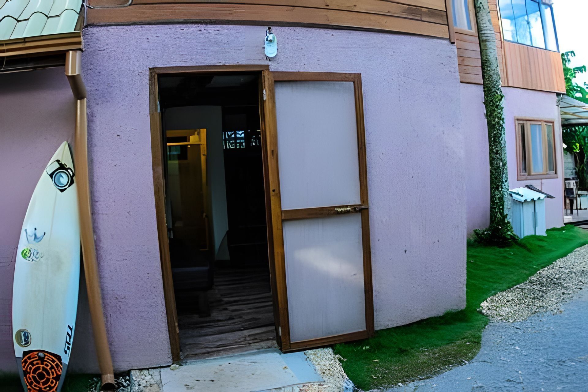 Un bâtiment avec une porte donnant sur un espace intérieur, une planche de surf appuyée contre le mur et un petit jardin avec de l’herbe et du gravier.