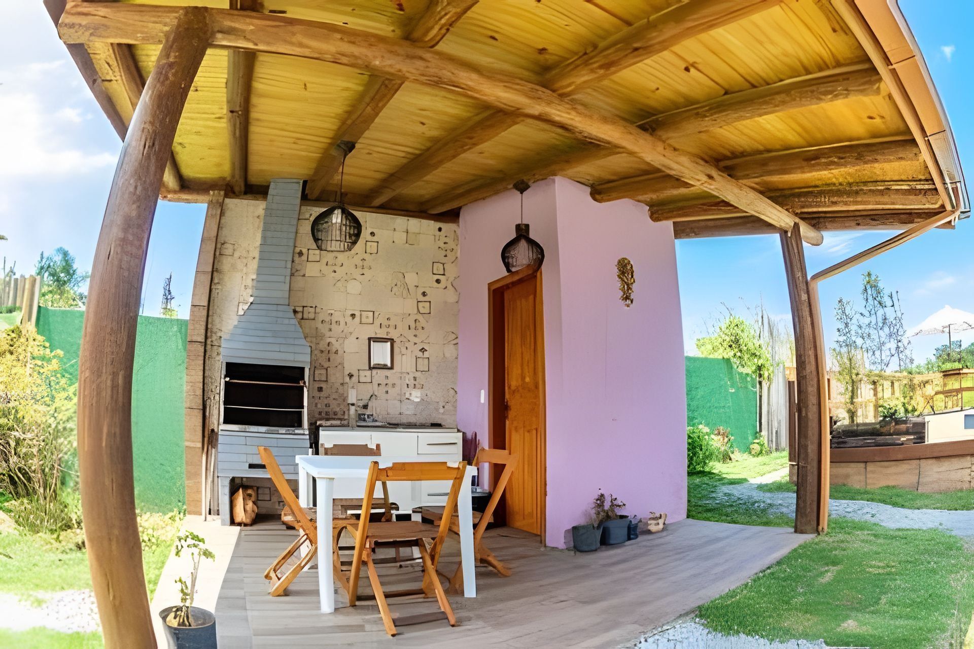 Un patio exterior con mesa y sillas de madera, una parrilla empotrada y una puerta que da a un jardín.