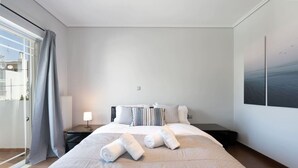 A hotel room with a large bed, a nightstand, a lamp, and a view of the sea through the window. - Luxe Bliss – 2BR, Glyfada (Glifada)