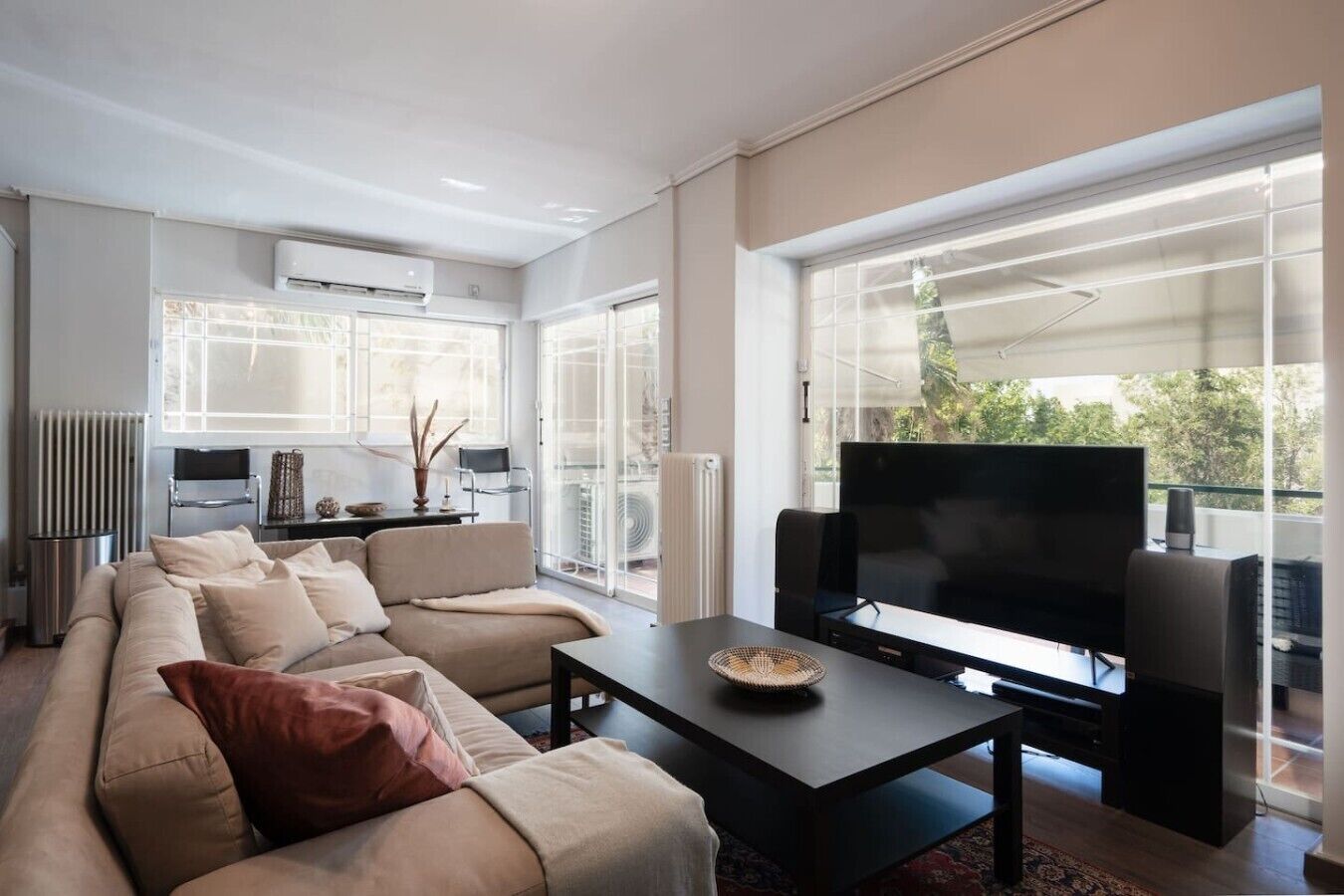 A modern living room with a sofa, a coffee table, and a flat-screen TV. There is a large window providing a view of the outdoors.